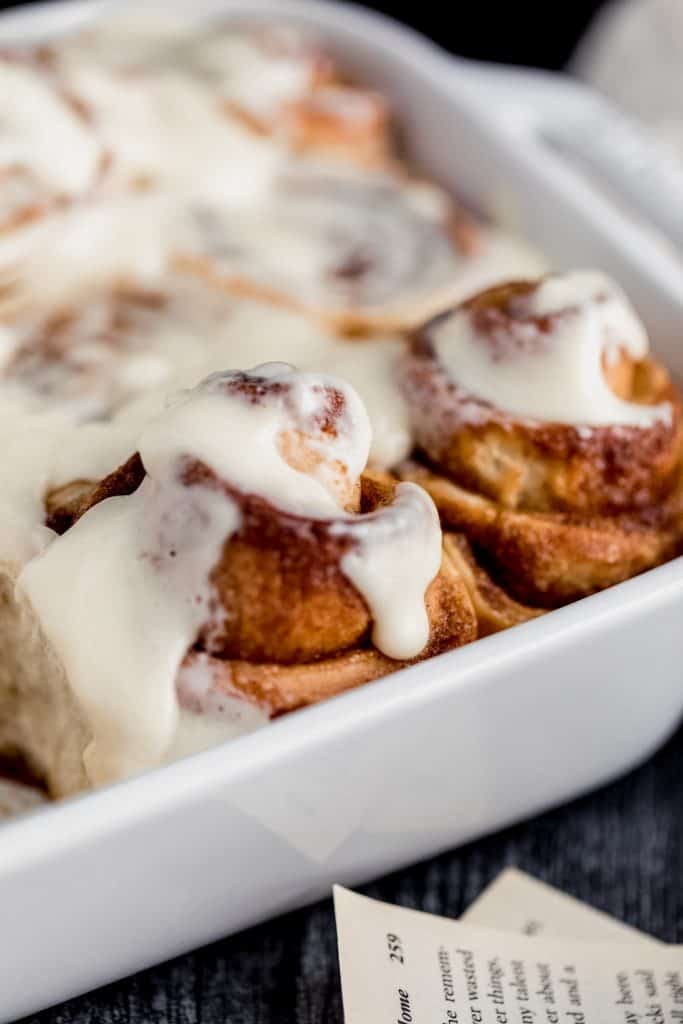 freshly baked and frosted cinnamon rolls in a white pan sitting on a dark weathered wood surface with a couple of book pages nearby
