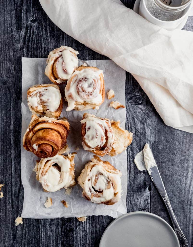 cinnamon rolls outside of the baking dish on parchment paper with cream cheese frosting on top of all but one of the rolls. a set of plates, a linen and a butter knife with frosting laying next to the pan