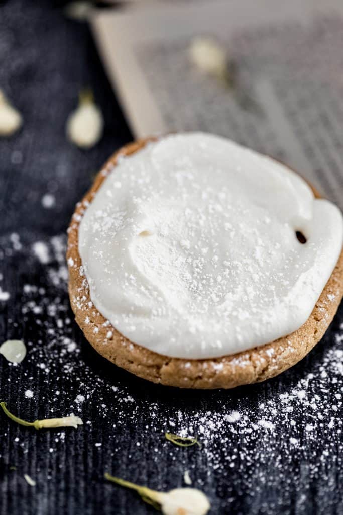 an open face oatmeal cream pie with all the filling on top, dusted in powdered sugar with dried white flowers sprinkled around on a dark weathered surface with a book page in the back corner