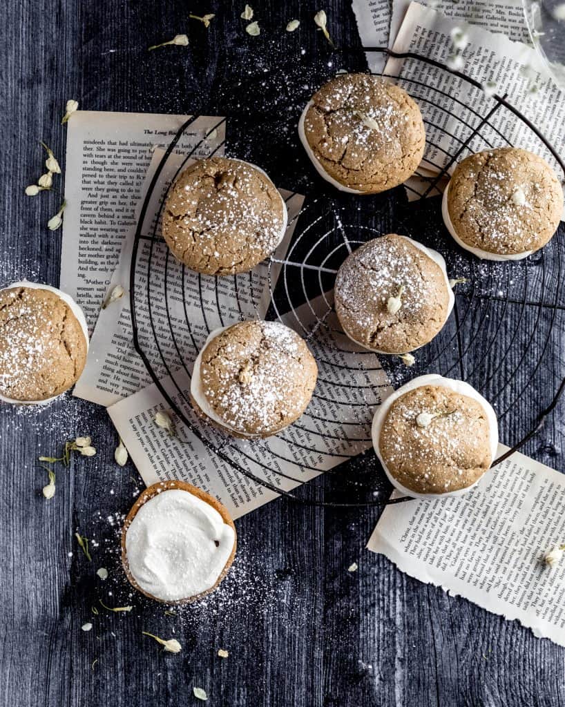 oatmeal cream pies on top of a circular cooling rack with book pages underneath dusted with powdered sugar and dried white flowers. one oatmeal cream pie is off to the side, open face so you can see all the cream