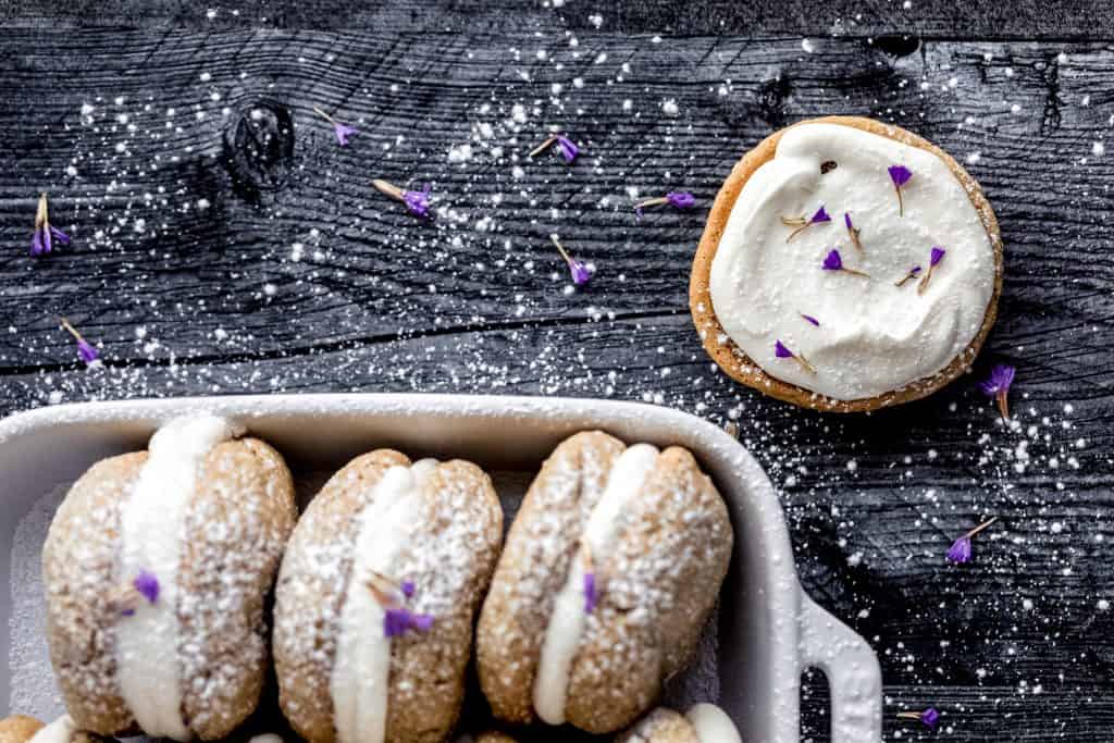 oatmeal cream pies lined up in a white baking dish with powdered sugar and lavender sprinkled on top. one open face cookie with cream filling on top in the upper right corner also sprinkled with powdered sugar and lavender