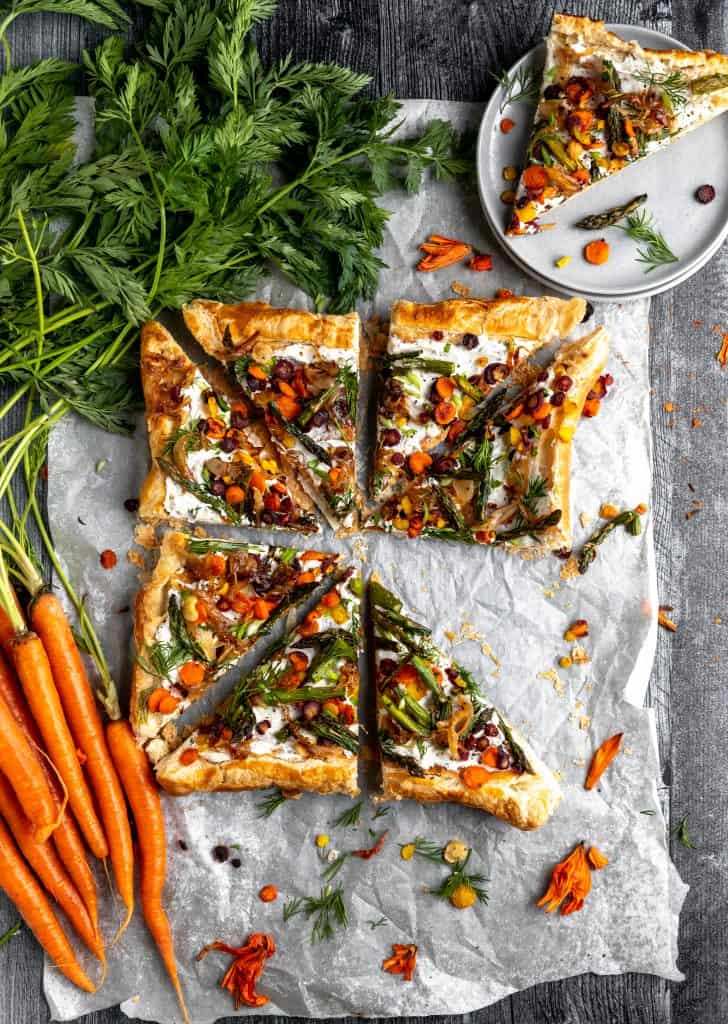 Asparagus tart with roasted veggies and carrots with green tops laying to the side. one triangle of tart missing out of the square