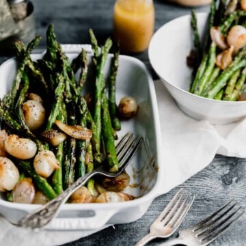 grilled asparagus sitting in a white baking dish topped with glazed shallots and sherry vinaigrette with one fork in the dish and 2 forks on the side. another plate of asparagus in the top right corner with the dressing to the left if it