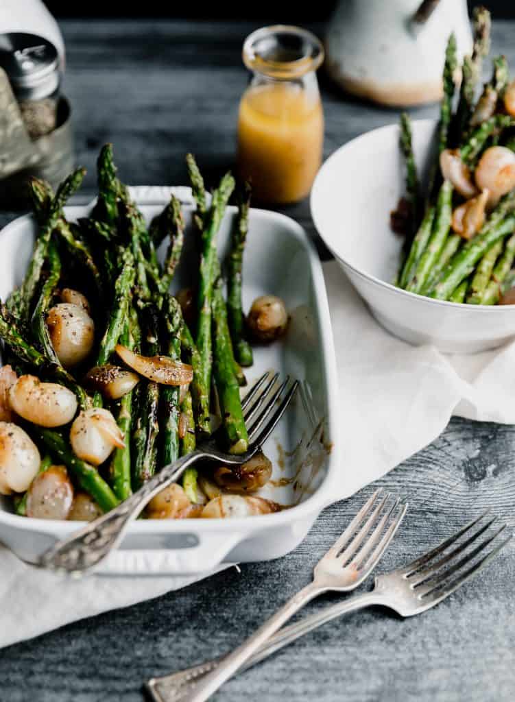 grilled asparagus sitting in a white baking dish topped with glazed shallots and sherry vinaigrette with one fork in the dish and 2 forks on the side. another plate of asparagus in the top right corner with the dressing to the left if it