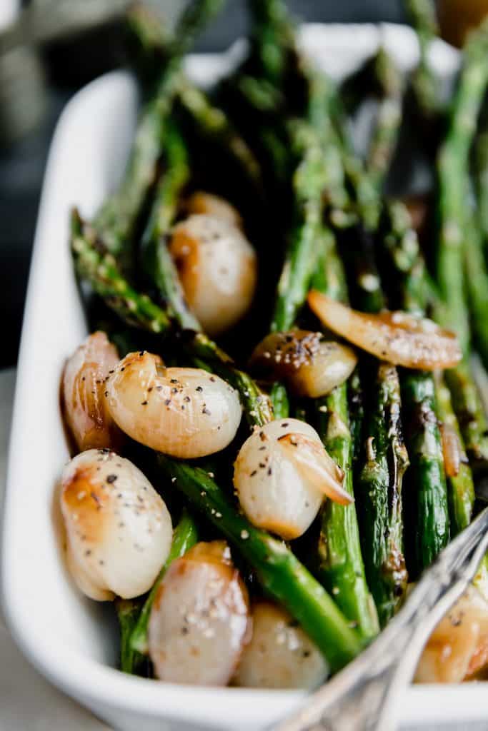 close up of grilled asparagus in a white baking dish with glazed shallots and sherry vinaigrette with a fork in the corner of the pan