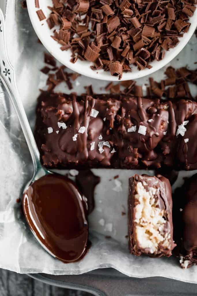 pecan joy candy bar on a tray with a spoon of melted chocolate, flaky sea salt, chocolate shavings and a cut candy bar on top of parchment paper