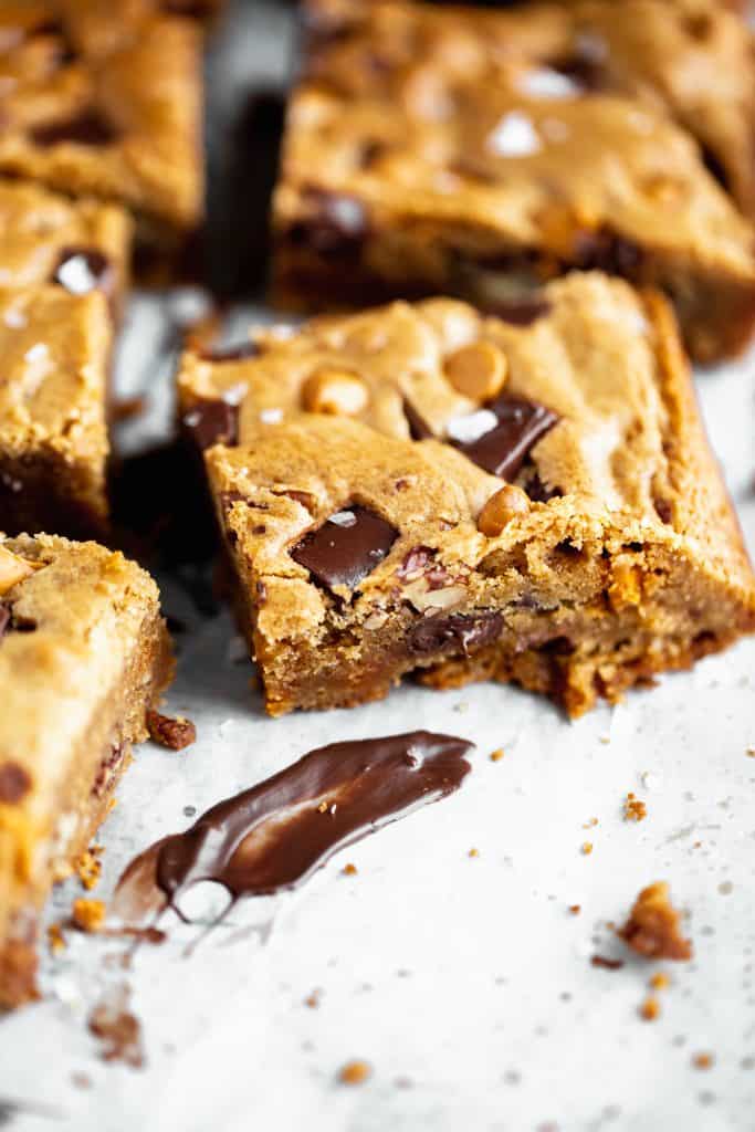 the best blondies with chocolate, butterscotch, toasted pecans and sea salt sitting on a white surface with a smear of melted chocolate in front of on of the pieces
