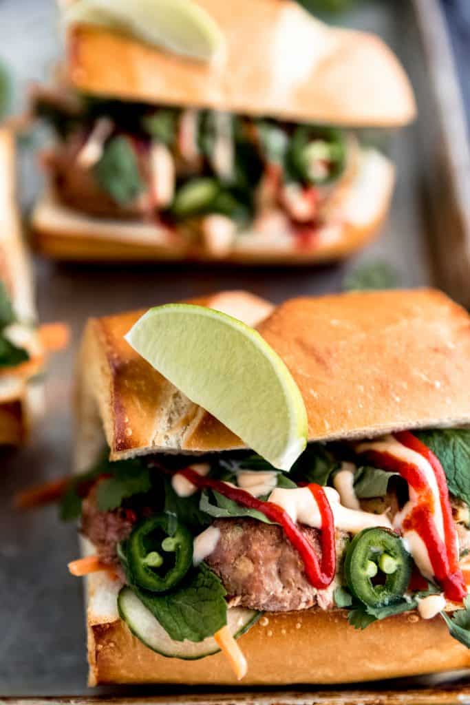 close up of the meatball banh mi with sriracha sauce, sweet chili sauce, lime wedge and various green herbs