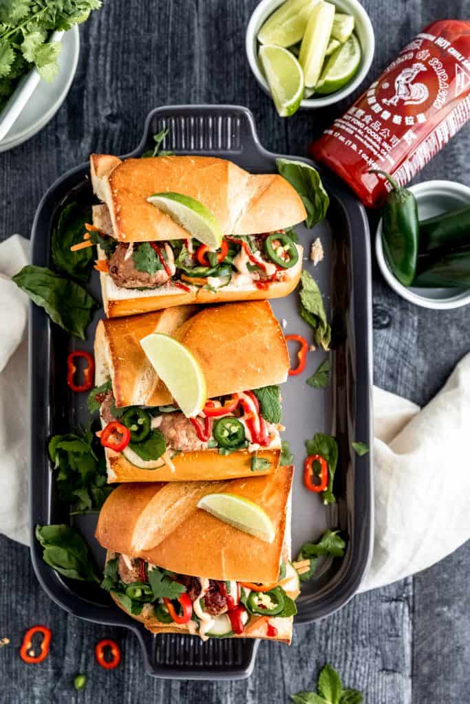 turkey meatball banh mi sandwiches on a grey platter on top of a dark weathered wood surface with a bottle of sriracha and various green toppings such as limes, mint, basil, cilantro and fresh jalapeno