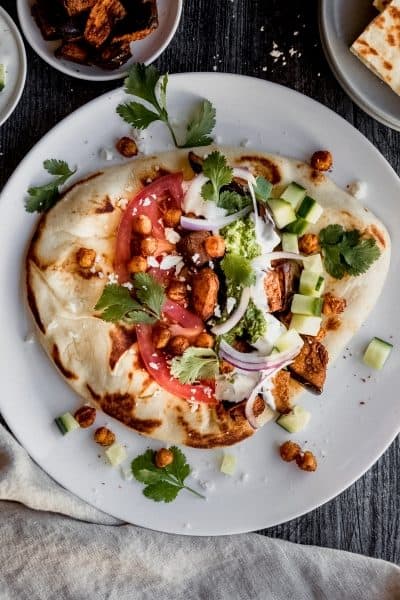 eggplant shawarma on naan with tzatziki sauce, schug, roasted garbanzo beans, cucumbers, tomatoes, red onion and cilantro on a white plate sitting on top of a dark weathered wood surface with a grey linen