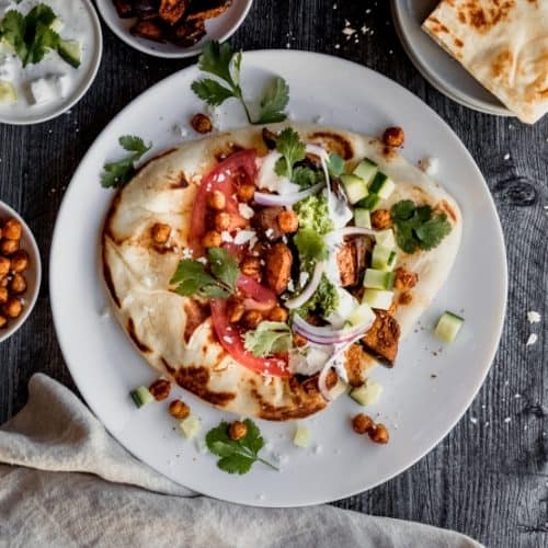 eggplant shawarma on naan with tzatziki sauce, schug, roasted garbanzo beans, cucumbers, tomatoes, red onion and cilantro on a white plate sitting on top of a dark weathered wood surface with a grey linen