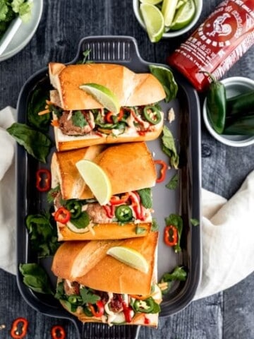 turkey meatball banh mi sandwiches on a grey platter on top of a dark weathered wood surface with a bottle of sriracha and various green toppings such as limes, mint, basil, cilantro and fresh jalapeno