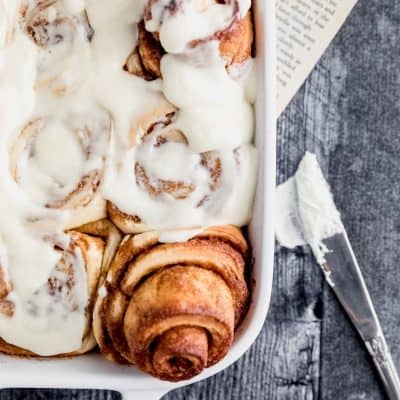 a white baking dish filled with ooey gooey cinnamon rolls with cream cheese icing. a knife to the side with icing on it and a book page underneath the pan sitting on a dark surface of weathered wood