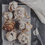ooey gooey cinnamon rolls spread out on top of white parchment paper with all but one roll topped with cream cheese frosting, a white linen and a butter knife with frosting next to it