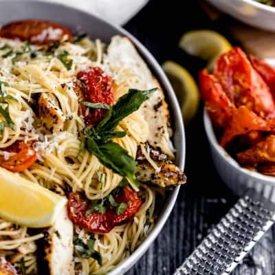 a bowl of garlic chicken pasta with a lemon wedge, grated asiago cheese, fresh basil and roasted tomatoes in it. A microplane grater off to the side with a bowl of roasted tomatoes and squeezed lemon wedges