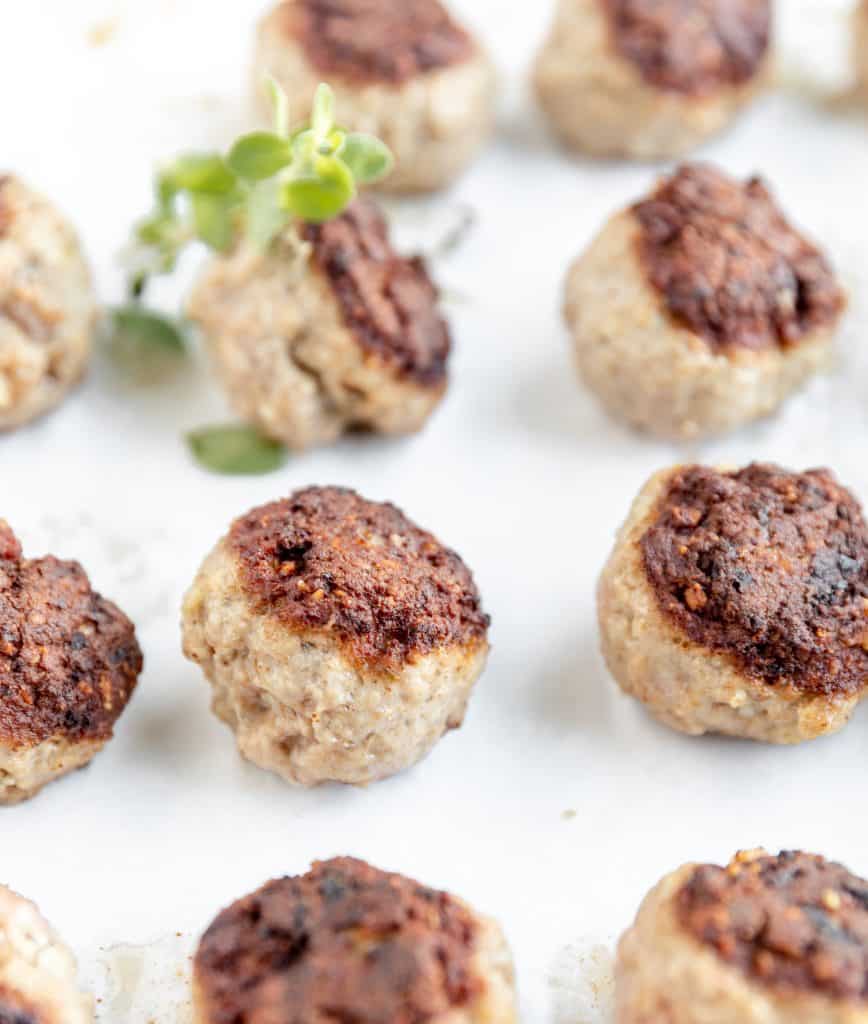 close up shot of seared meatballs sitting on a tray with parchment paper underneath them and some fresh oregano