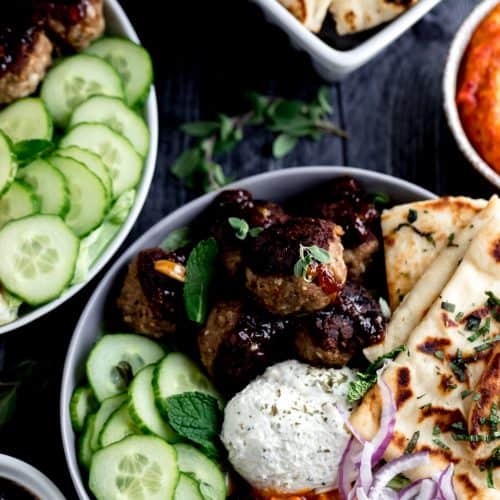 2 mediterranean bowls with meatballs, cucumbers, garlic cheese spread, naan, red onion, chutney and mint leaves in a grey and white bow with a bowl of cherry pepper hummus on the side on a dark surface