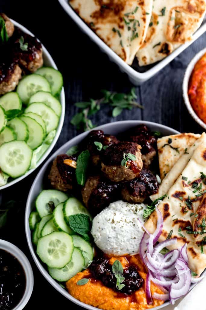 2 mediterranean bowls with meatballs, cucumbers, garlic cheese spread, naan, red onion, chutney and mint leaves in a grey and white bow with a bowl of cherry pepper hummus on the side on a dark surface