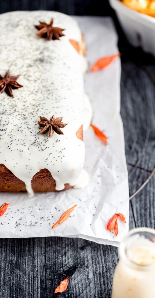 a whole loaf of orange poppy seed bread with yogurt glaze sitting on top of white parchment paper topped with whole star anise and poppy seed. orange dried flowers scattered on the parchment paper