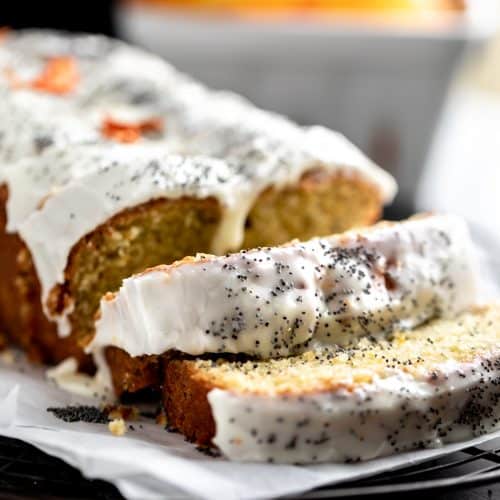 Orange Poppy Seed Bread with an orange yogurt glaze sitting on top of parchment paper on a cooling rack. 2 slices and the rest of the loaf is whole. topped with poppy seeds and dried orange flowers