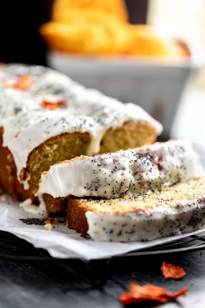 Orange Poppy Seed Bread with an orange yogurt glaze sitting on top of parchment paper on a cooling rack. 2 slices and the rest of the loaf is whole. topped with poppy seeds and dried orange flowers