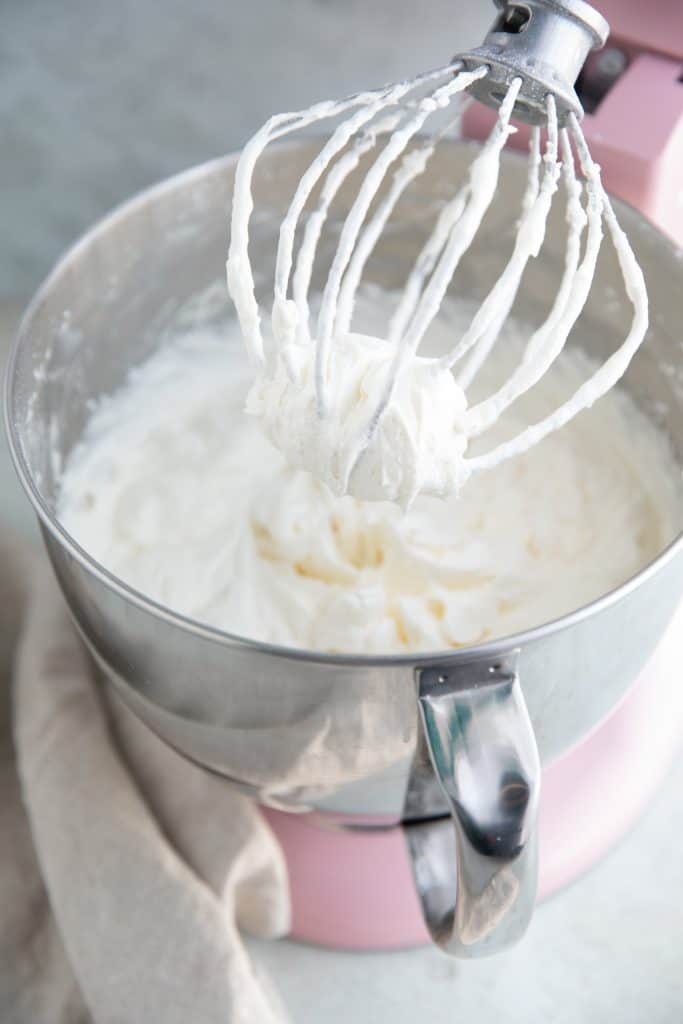 almond frosting in a pink kitchen aid mixer with a whisk attachment lifted up out of the bowl with the frosting on it