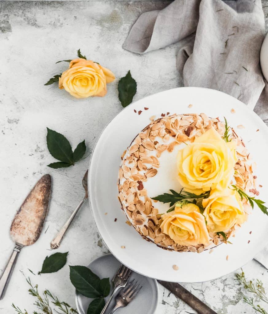 top view of almond carrot cake with toasted almonds and yellow roses on top surrounded by a linen, cake spatula and roses with leaves