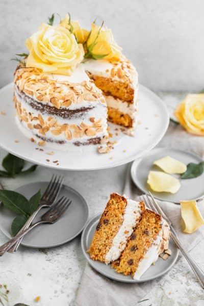 almond carrot cake on a cake stand with one slice cut out of it sitting on a plate in front of the full cake. yellow roses on top of the cake as well as scattered around the counter top. toasted almonds on the outside of the cake