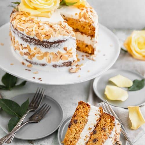 almond carrot cake on a cake stand with one slice cut out of it sitting on a plate in front of the full cake. yellow roses on top of the cake as well as scattered around the counter top. toasted almonds on the outside of the cake