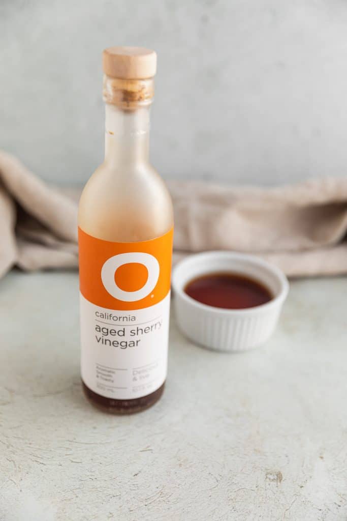 a bottle of sherry vinegar with a white ramekin next to it filled with sherry vinegar sitting on top of a light surface with a linen cloth behind them