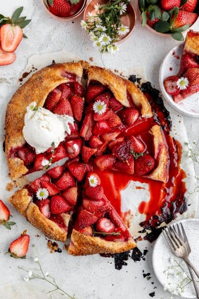 strawberry galette cut into triangles with one slice sitting on a white speckled plate and a scoop of honey vanilla ice cream on top of the whole tart garnished with white daisies