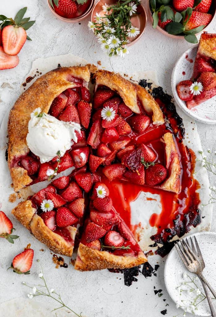 strawberry galette cut into triangles with one slice sitting on a white speckled plate and a scoop of honey vanilla ice cream on top of the whole tart garnished with white daisies