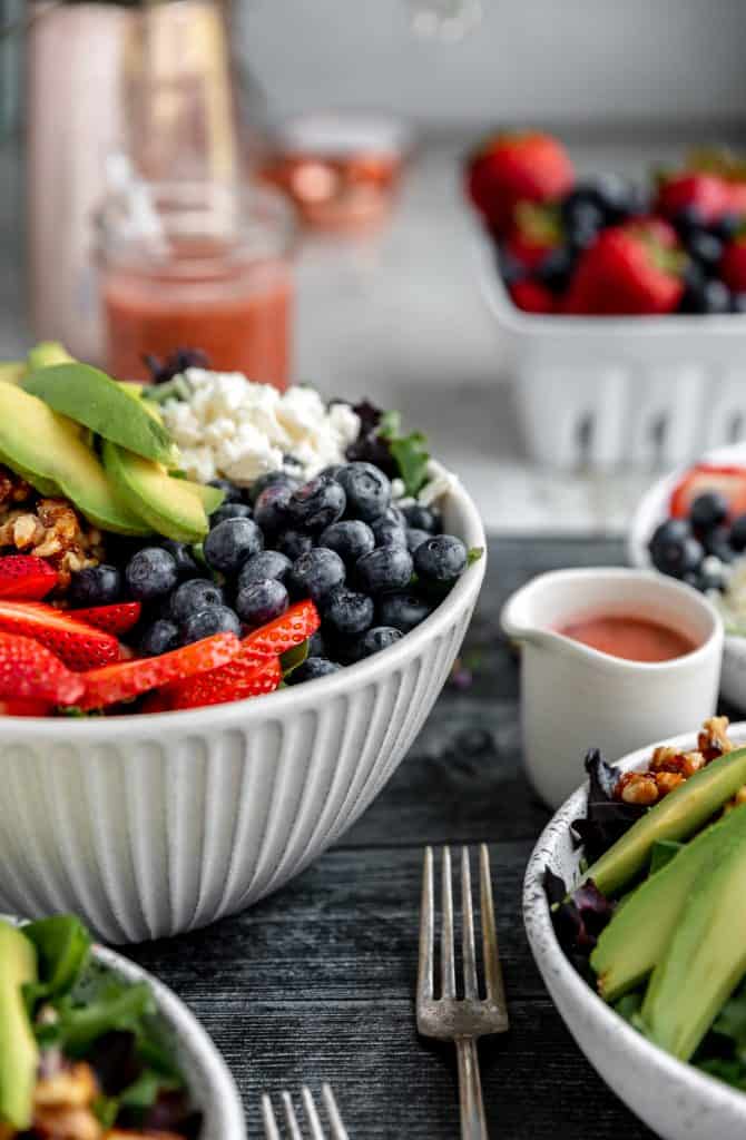 Multiple bowls of strawberry summer salad filled with strawberries, blueberries, avocado, feta cheese and candied walnuts. strawberry vinaigrette in a jar