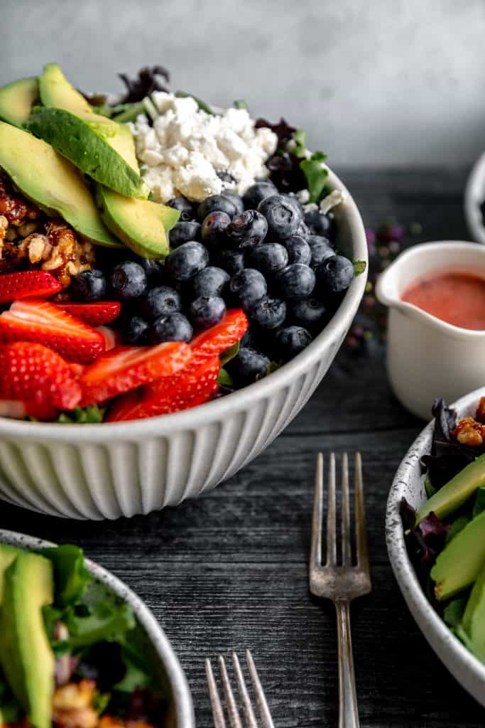 a large bowl of strawberry summer salad filled with strawberries, blueberries, avocado, feta, candied walnuts, mixed greens and a container of strawberry vinaigrette