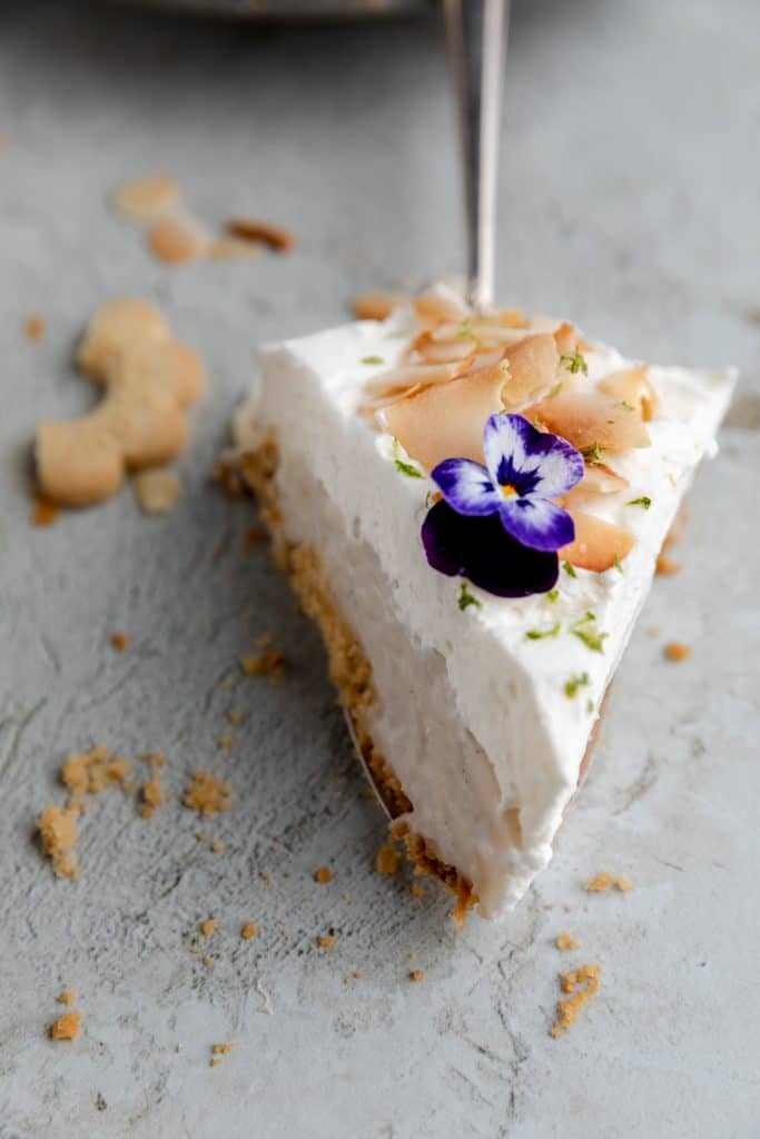 a slice of coconut cream pie on a pie server topped with toasted coconut flakes and a purple flower