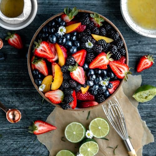summer fruit in a bowl with the dressing, some limes, strawberries and a whisk to the side