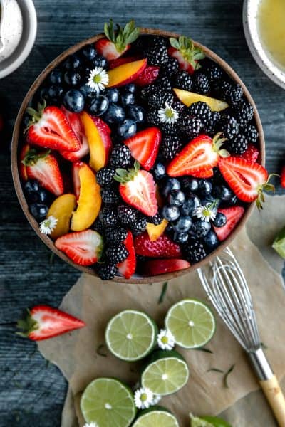 summer fruit salad mixed with lime ginger dressing with limes and a whisk to the side