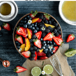 summer fruit salad in a bowl with honey and the lime dressing to the side with some cut limes and strawberries lying around the bowl