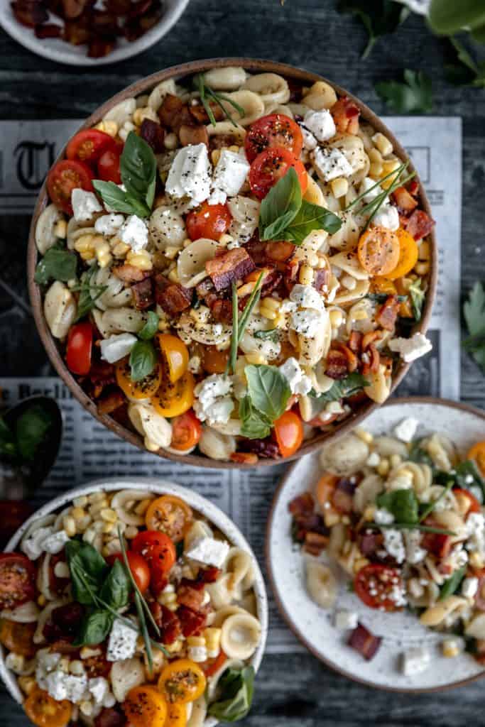 summer pasta salad with corn, bacon, tomatoes and feta cheese in 2 bowls and a plate