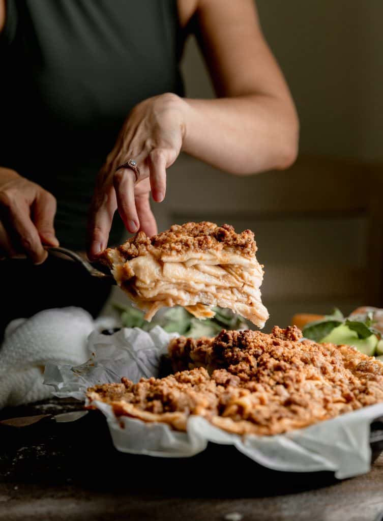 a woman lifting a slice of apple pie out of the cast iron skillet to be served