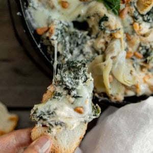 Spinach Artichoke dip in a cast iron skillet with a piece of toast being dipped into it.