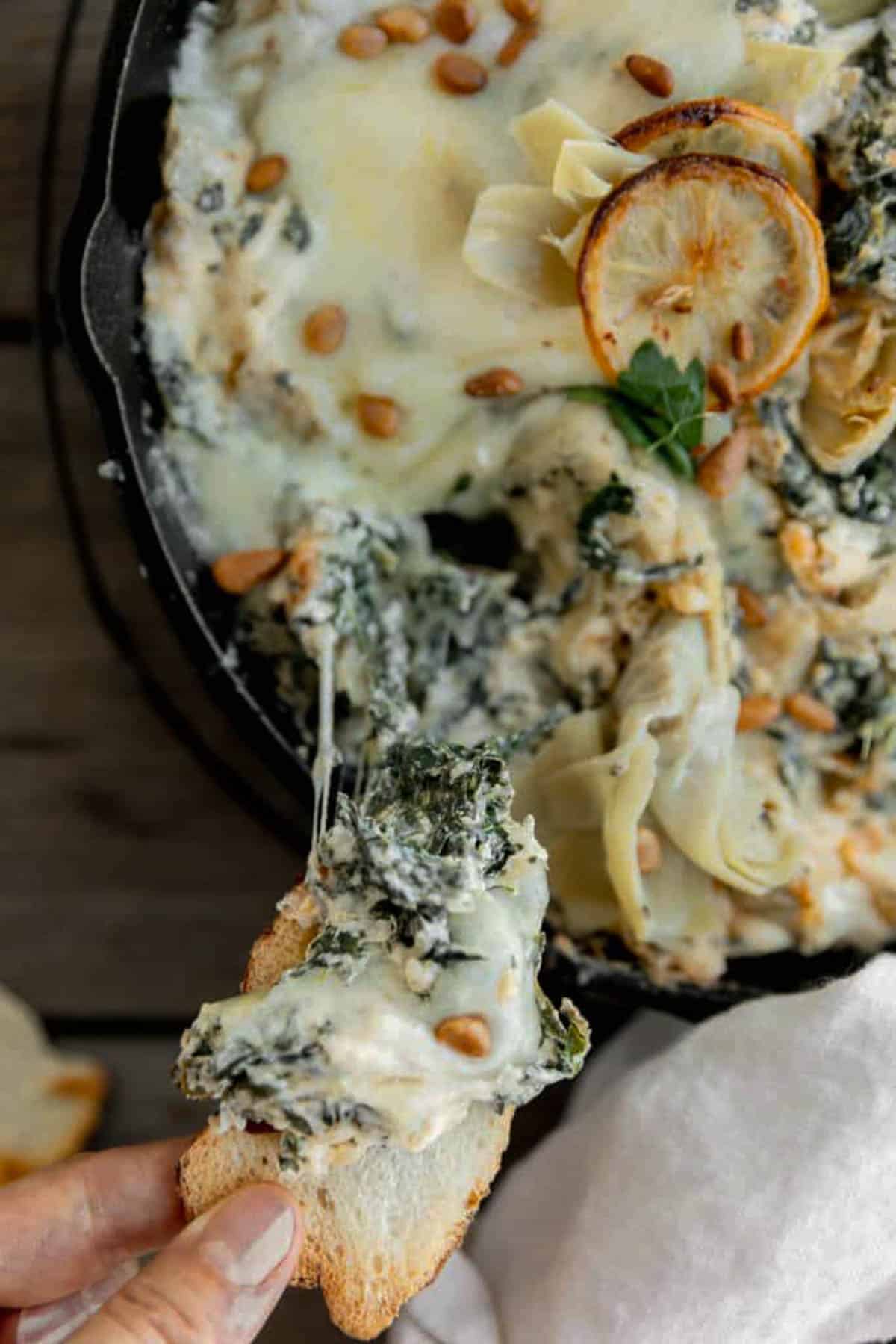 Spinach Artichoke dip in a cast iron skillet with a piece of toast being dipped into it. 