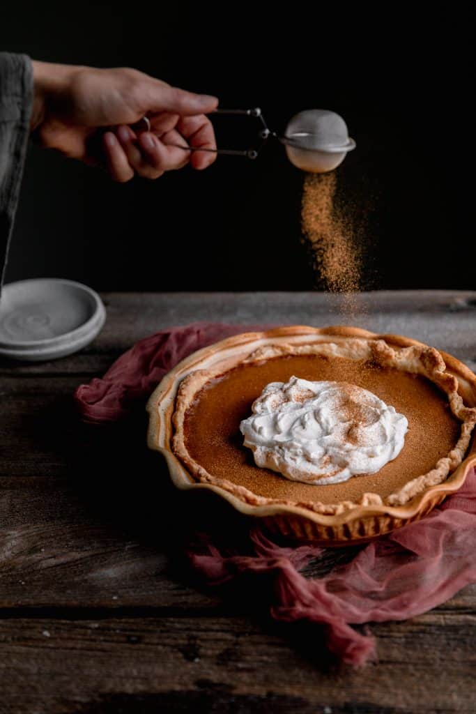 dusting cinnamon on top of pumpkin pie that's been topped with whipped cream