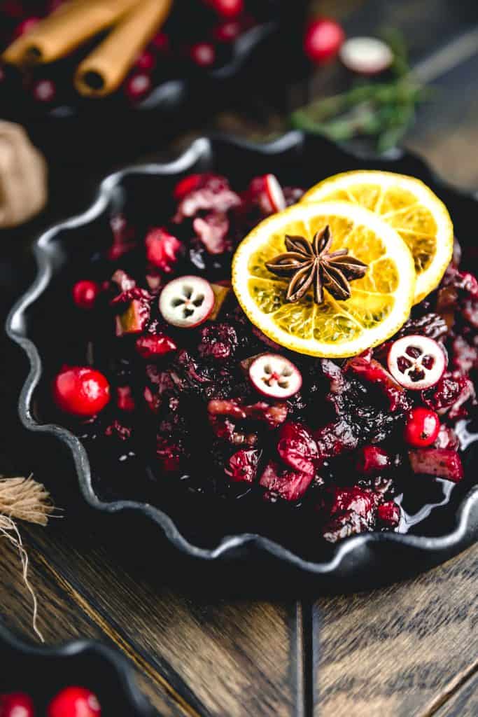A black bowl filled with cranberry orange sauce topped with orange slices, sliced cranberries and star anise on a wooden table.