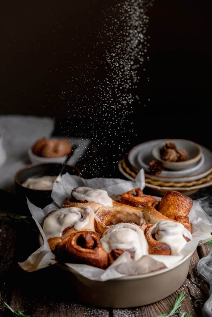 a pan of gingerbread cinnamon rolls topped with eggnog frosted being dusted with powdered sugar
