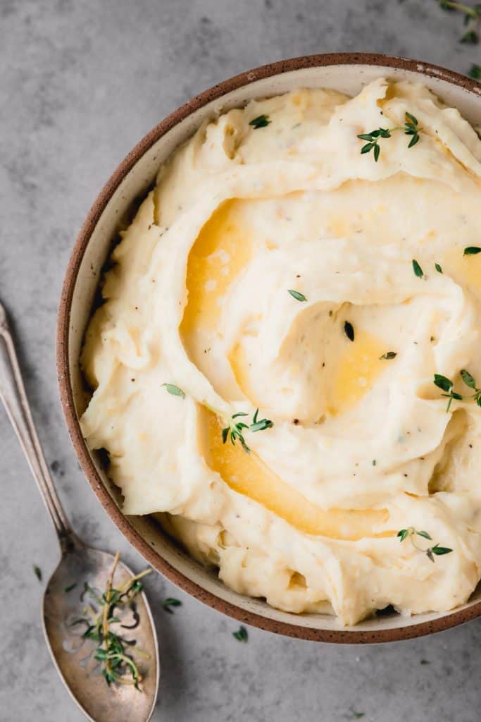 Boursin mashed potatoes swirled with butter and thyme in a white bowl with a brown rim. A serving spoon laying to the left side of the bowl. 