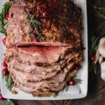 Standing Rib Roast partially sliced on a platter with fresh rosemary and cranberries.