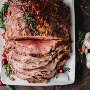 Standing Rib Roast partially sliced on a platter with fresh rosemary and cranberries.
