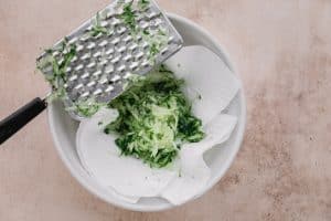 shredding cucumber into a paper towel lined bowl with a grater