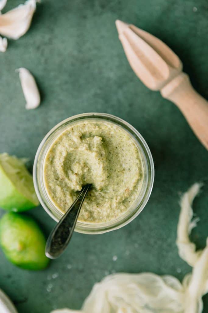 creamy taco truck green salsa in a jar with a spoon in it. limes lying to the side with a juicer and garlic cloves